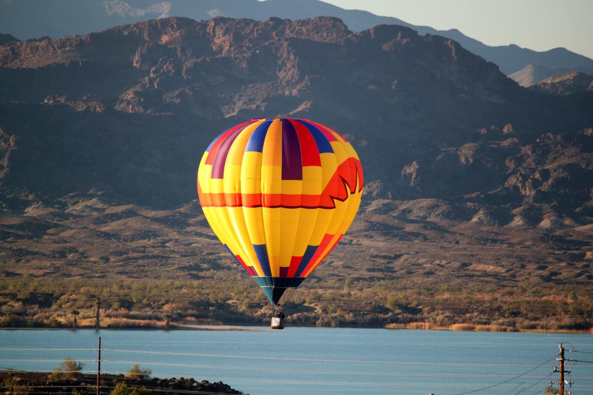 Hot air balloon drifting over open water below desert mountains